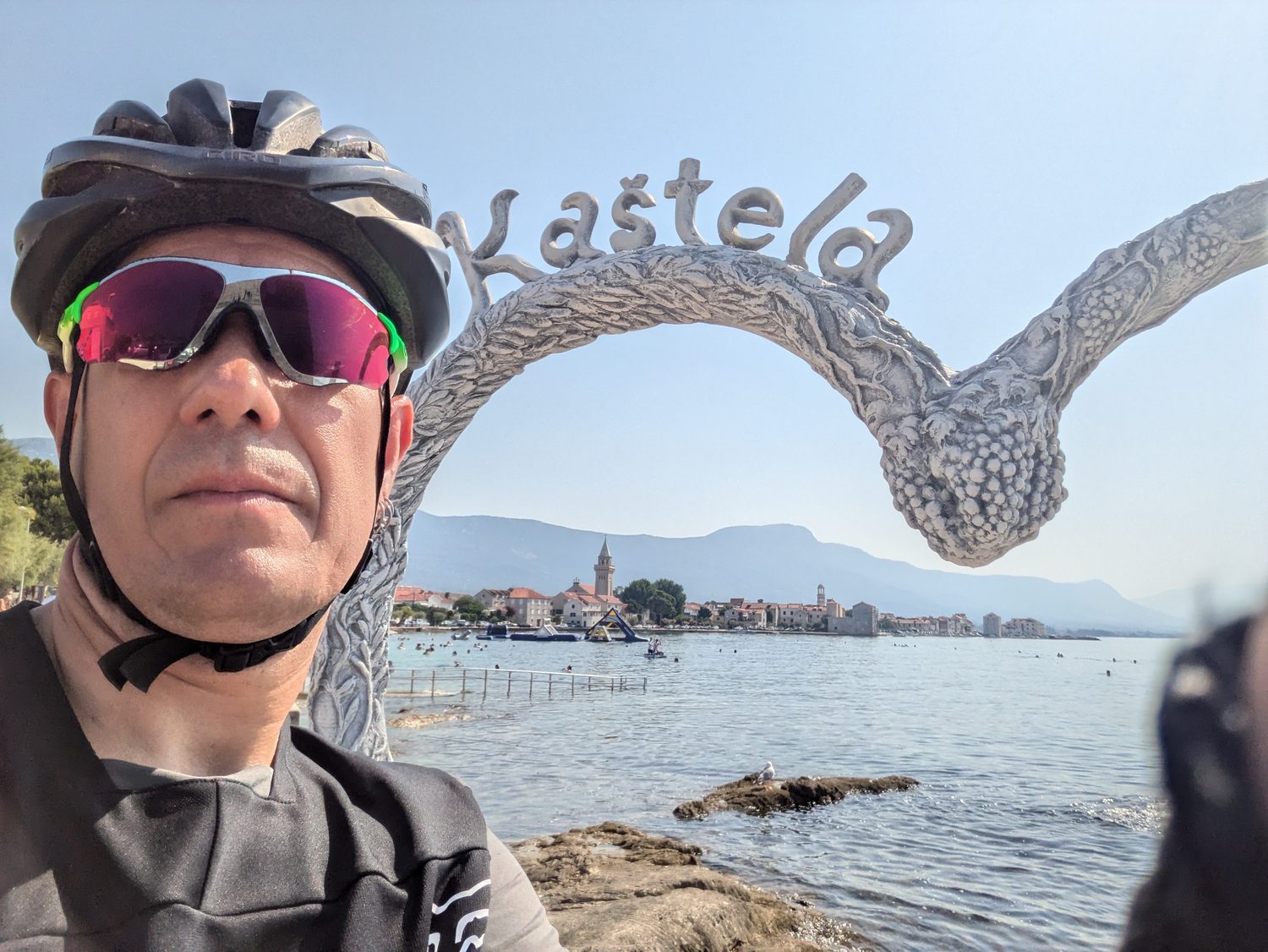 Fietser met helm en zonnebril voor de Kaštela-boog aan de kust, met op de achtergrond een dorp met kerktoren en de baai