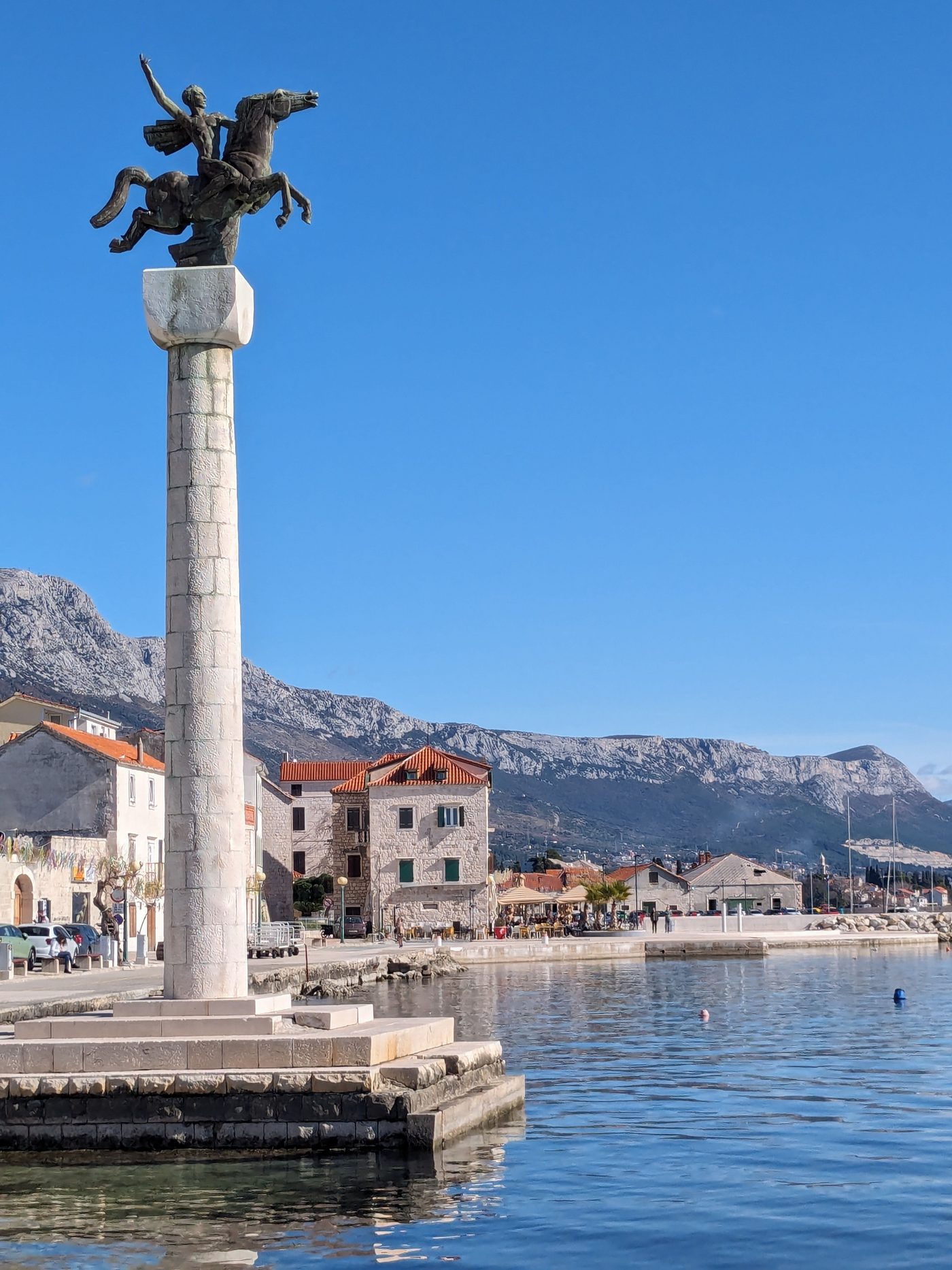 Bronzen ruiterstandbeeld op een hoge stenen zuil aan de haven van Kaštel Stari, met besneeuwde bergtoppen van de Kozjak op de achtergrond